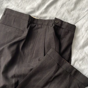 Vintage 80-tal Svarta randiga kostymbyxor - Svarta kostymbyxor med tunna vertikala ränder. Byxorna har klassisk passform med pressveck, hällor för bälte och knäppning fram. Tillverkade i ett ganska lätt polyestertyg. Mått: midja: 80cm, innerben: 84cm, Byxöppning: 23cm (halva) Midjehöjd 33cm. Hög till mellanhög midja. 