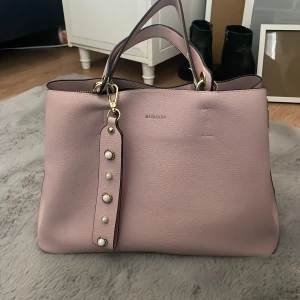 Ljusrosa handväska från Marhen.J - Snygg ljusrosa handväska från Marhen.J med pärldetaljer på axelremmen och guldfärgade metalldelar. Väskan har flera fack, dragkedja och magnetlås. Tillverkad i slätt syntetmaterial och har både handtag och axelrem för olika stilar.