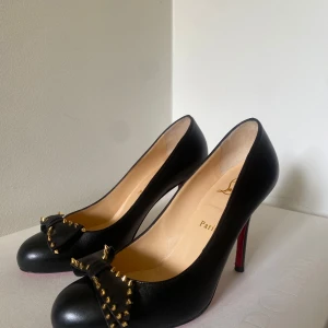 Svarta Louboutin pumps med nitar - Svarta pumps i skinn från Christian Louboutin med klassisk röd sula. Skorna har rund tå, hög klack och är dekorerade med en rosett framtill med guldiga nitar för en edgy vibe. Perfekta för dig som vill sticka ut med lyxig och cool stil.