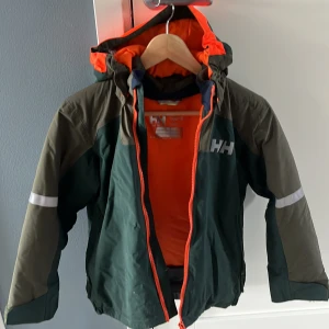 Grön skidjacka barn från Helly Hansen - Cool grön och olivfärgad vindjacka från Helly Hansen med orange detaljer och huva. Jackan har reflekterande band på ärmarna, dragkedja framtill och logga på bröstet. Perfekt för vinterdagar och utomhusaktiviteter. Använd 2 ggr. Storlek 140. Katt finns i hemmet.