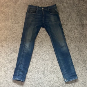 Replay Anbass - Säljer ett par Replay Anbass Hyperflex jeans i blå tvätt med snygga slitningar. Modellen är slim fit med klassiska fem fickor och coola detaljer på myntfickan. Jeansen är stretchiga och sitter slim men bekvämt. Perfekta för dig som gillar en modern look.