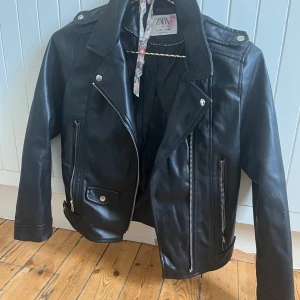 Svart skinnjacka från Zara - Snygg svart skinnjacka från Zara med klassisk bikerstil. Jackan har silvriga dragkedjor, tryckknappar och coola detaljer vid axlarna. Fodrad insida för extra komfort. Perfekt för dig som vill ha en edgy look till vardags.
