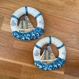 Dekoration Väggprydnader - Säljer två dekorativa väggprydnader i keramik med marint tema, föreställande segelbåtar omgivna av livbojar och blå vågor. 