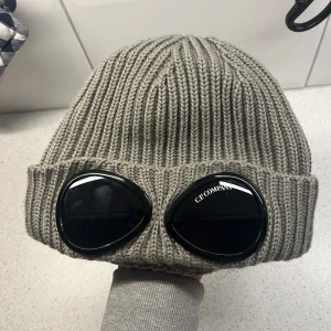 Grå ribbstickad mössa C.P. Company - ÄKTA KÖPT I BUTIK. HELT OANVÄND!                            PRIS KAN DISKUTERAS🔥                                             Snygg grå ribbstickad mössa från C.P. Company med ikoniska svarta goggles framtill. Mössan har uppvikt kant och är tillverkad i mjukt stickat material som håller dig varm och ger en cool streetstyle-vibe.