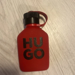 Hugo Boss röd parfymflaska ca 68/75 ml kvar - Cool parfym från Hugo Boss i en matt röd flaska med svart skruvkork och praktisk röd rem. Flaskan har tydlig svart text 'HUGO' på framsidan och en modern, stilren design som sticker ut i badrumshyllan.