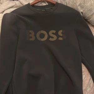 Svart sweatshirt från BOSS med logga - Svart sweatshirt från BOSS med guldig logga framtill i coolt randigt tryck. Klassisk rund halsringning och långa ärmar. Perfekt för dig som gillar stilrena och trendiga plagg med premiumkänsla.