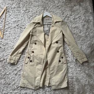 Beige trenchcoat från Stefanel - Snygg beige trenchcoat från Stefanel med klassisk dubbelknäppning, bred krage och bälte i midjan. Jackan har långa ärmar och fickor framtill. Perfekt för dig som vill ha en stilren och tidlös look.