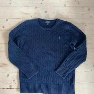 Marinblå kabelstickad tröja Polo Ralph Lauren - Kabelstickad marinblå tröja från Polo Ralph Lauren med rund halsringning och grönt broderat logomärke på bröstet. Tröjan har långa ärmar och breda ribbade muddar vid ärmslut och nederkant. Perfekt för en stilren och klassisk look.