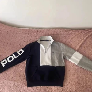 Colorblock half zip tröja Polo Ralph Lauren - Snygg colorblock tröja från Polo Ralph Lauren med half zip och stor POLO-logga på ärmen. Färgerna är marinblå, grå och vit. Klassisk krage, långärmad och med ficka framtill. Perfekt för dig som gillar sportig och stilren look.