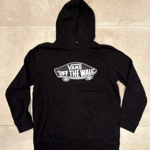 Svart Vans hoodie med tryck - Svart hoodie från Vans med stort vitt 'Off The Wall'-tryck på bröstet. Klassisk känguruficka framtill och huva. Passar som M