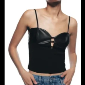 Svart korsett-topp med cut-outs - Snygg svart korsett-topp med tunna axelband och coola cut-outs framtill. Toppen har glansiga kupor i skinnimitation och croppad passform som ger en edgy vibe. Perfekt att styla med jeans eller kjol för en trendig look.