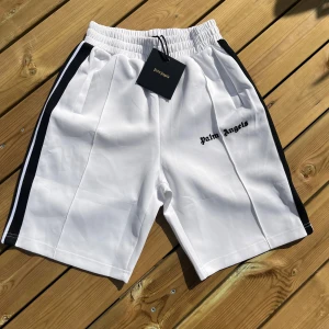 Palm Angels vita shorts med svarta ränder - Snygga vita shorts från Palm Angels med svarta sidor och broderad logga framtill. De har elastisk midja och sportig look, perfekta för en chill och trendig stil. Materialet är mjukt och känns lyxigt, med klassiska sidofickor och raka ben.
