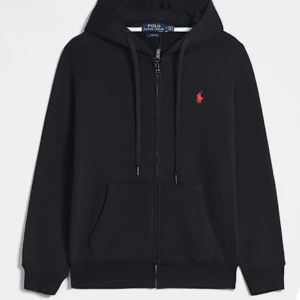 Svart hoodie från Polo Ralph Lauren - Svart hoodie från Polo Ralph Lauren med röd broderad logga på bröstet. Tröjan har huva med snörning, dragkedja framtill och känguruficka. Tillverkad i mjuk bomull och har långa ärmar. Perfekt för en chill och stilren look.