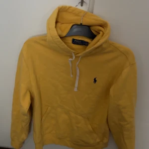 hoodie från Polo Ralph Lauren - Gul hoodie från Polo Ralph Lauren med klassisk blå logga broderad på bröstet. Tröjan har huva med vit snörning, känguruficka och ribbade muddar. Perfekt för dig som gillar en färgstark och chill stil.