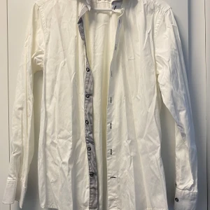 Vit skjorta från Stenströms med ränder - Vit långärmad skjorta från Stenströms med smal passform. Snygga svarta och vita ränder på insidan av kragen och knappslån, samt kontrastknappar i svart och en röd detalj på manschetten. Tillverkad i tvåfaldig superbomull för extra mjuk känsla.