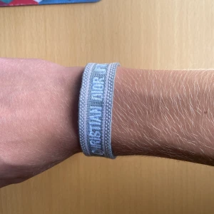 Grå Christian Dior J'Adior armband - Snyggt grått armband från Christian Dior med texten 'J'Adior' broderat i vitt. Armbandet är gjort i ett mjukt tygmaterial och har två stilrena tofsar i ändarna. Perfekt accessoar för att lyfta din outfit med en lyxig touch.
