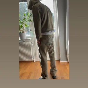 Syna-world tracksuit - Säljer ett par beige mjukisbyxor med fett graffiti-inspirerat tryck på baksidan. Byxorna har normal passform, elastisk midja med snörning och är gjorda i mjukt bomullsmaterial. Perfekt för chill eller streetwear-stil. Snygga detaljer och skön vibe.