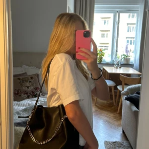 Svart axelväska med kedjedetalj från Núnoo - Säljer en svart axelväska från Núnoo i slätt skinn med justerbart axelband och en silverkedja framtill. Väskan är rymlig med magnetlås och innerfack med dragkedja. Perfekt för dig som vill ha en väska till skolan eller annat! Väskan har inga defekter och är i nyskick! Pris kan diskuteras i snabb affär😊