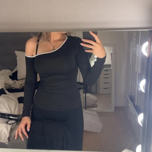 Svart oneshoulder topp med vit kant - Snygg svart långärmad topp med oneshoulder-design och vit kant längs halsringningen. Tight passform som framhäver figuren och ger en modern vibe. Perfekt för dig som gillar stilrena plagg med en twist.