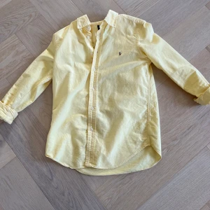 Gul skjorta från Ralph Lauren - Snygg ljusgul skjorta från Ralph Lauren med klassisk krage och broderad logga på bröstet. Skjortan har långa ärmar, knäppning framtill och är gjord i mjuk bomull. Perfekt för dig som gillar stilrena och fräscha plagg.