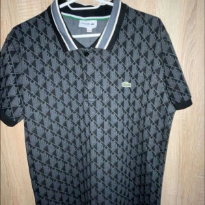 Svart mönstrad t-shirt från Lacoste - Svart t-shirt från Lacoste med grått geometriskt mönster över hela plagget. Klassisk krage med vita och grå ränder, grönt Lacoste-märke på bröstet och gröna/vita ränder längs axlarna. Kortärmad och tillverkad i mjuk bomull. 