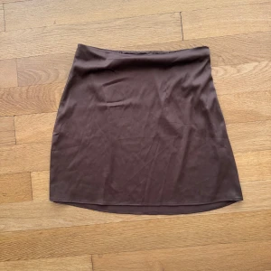 Brun satinkjol - Nelly by Bianca - En enkel och stilren brun kortkjol med rak passform. Kjolen är gjord i ett mjukt och slätt material som känns skönt mot huden. Perfekt att matcha med en cool topp eller hoodie för en avslappnad look.