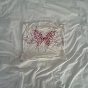 Vit tubtopp med röd fjäril - Vit tubtopp med en stor röd fjäril och dekorativa mönster framtill. Toppen är axelbandslös och har en enkel, rak passform. Perfekt för dig som vill sticka ut med ett snyggt tryck och en clean look.