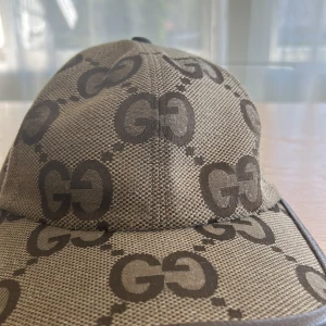 Beige keps från Gucci med GG-mönster - Snygg beige keps från Gucci med det klassiska GG-logomönstret i mörkbrunt. Kepsen har justerbart spänne bak i brunt och är tillverkad i ett slitstarkt bomullstyg. Perfekt för dig som vill ha en exklusiv och trendig accessoar.