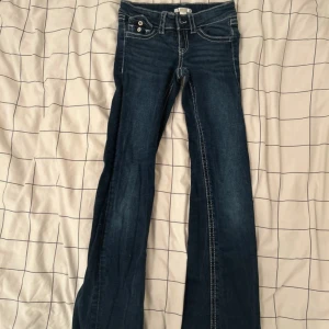 Blå bootcut flare jeans från Gina Tricot - Snygga blå jeans från Gina Tricot med bootcut passform och kontrastsydda sömmar. Klassisk femficksmodell med knappar och dragkedja fram samt fickor med lock och knapp bak. Perfekta för dig som gillar retrovibbar och markerade detaljer.