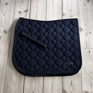 Eskadron schabrak - Snyggt, svart schabrak från Eskadron i quiltad design. Perfekt för dig som gillar hästar och vill ha en stilren look på ridturen. Tillverkat i 100% polyester, mjukt och slitstarkt. Passar både träning och tävling. Lite hår på insidan, annars fint skick.Använt en gång för typ 2 år sen. Mörkblått med lite halkskydd, dressyrschbrak som jag köpte för 800:- på rea så kostar mer än det i vanligt fall. Inga som helst skador, bara lite skitig men tvättar den såklart!  Storlek ponny/cob