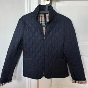 Mörkblå quiltad jacka  Burberry, strl S - Snygg mörkblå quiltad jacka från Burberry med klassiskt rutigt foder. Jackan har dragkedja framtill, två fickor och ståkrage. Storlek S