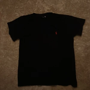 Mörkblå t-shirt från Ralph Lauren - Mörkblå t-shirt från Ralph Lauren med klassisk rund hals och korta ärmar i storlek M. På bröstet finns den ikoniska röda Polo-loggan broderad. T-shirten är gjord i mjuk bomull och har en clean, stilren look som funkar till allt.