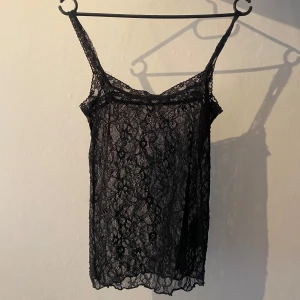 Svart spetsig linnetopp från Nelly  - Supersnygg svart linnetopp från Nelly i transparent spets med blommigt mönster. Tunt tyg och smala axelband ger en trendig look. Perfekt att styla lager på lager eller med ett par jeans för en cool vibe.