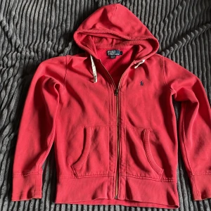 Röd hoodie från Polo Ralph Lauren - Snygg röd hoodie från Polo Ralph Lauren med klassisk blå logga broderad på bröstet. Tröjan har huva med vita snören, dragkedja framtill och två fickor. Tillverkad i mjuk bomull och polyester, perfekt för chill dagar. Långärmad och med ribbade muddar för extra komfort.