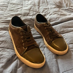Bruna sneakers från Lanvin i mocka - Snygga bruna sneakers från Lanvin med ovandel i mjuk mocka och glansig tå i mörkbrunt lack. Skorna har rund tå, snörning och vit platt sula. Perfekt för dig som gillar stilrena och exklusiva detaljer. Lanvin-logga på sulan. Riktigt schyssta till jeans eller chinos.