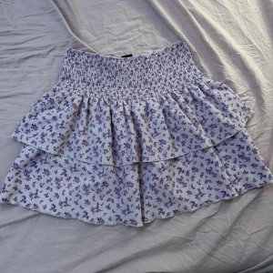 Ljuslila blommig volangkjol Gina Tricot - Supersöt kort kjol från Gina Tricot i ljuslila med små blommor. Kjolen har två lager volanger och en smockad midja som sitter bekvämt. Perfekt för dig som gillar romantisk och feminin stil. Materialet är mjukt och lätt, perfekt för varma dagar.