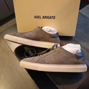 Grå mocka sneakers Axel Arigato - Snygga grå sneakers från Axel Arigato i mjuk mocka med rund tå och platt vit sula. Skorna har klassisk snörning och stilren design som passar till jeans eller chinos. Perfekta för dig som gillar minimalistisk och modern stil.