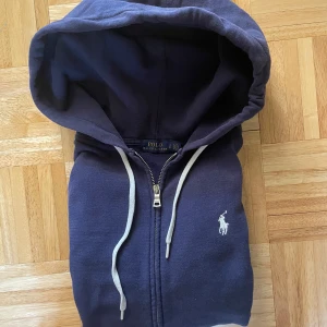 Mörkblå hoodie från Polo Ralph Lauren - Snygg mörkblå hoodie från Polo Ralph Lauren med vit broderad logga på bröstet. Tröjan har huva med vita snören och dragkedja framtill. Perfekt för chill dagar och har en klassisk streetstil. Materialet är mjuk bomull som känns skönt mot huden. OBS! Den är liten i storleken