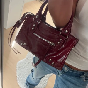 Vinröd handväska i skinn med dragkedja - Snygg vinröd handväska i glansigt skinn med silverfärgade detaljer och dragkedja framtill. Väskan har två handtag, dekorativa nitar och extra remmar som ger en cool vibe. Insidan har fack med dragkedja för småsaker. Perfekt storlek för det viktigaste.