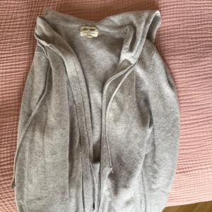 Grå hoodie i kashmir från Soft Goat - Mjuk och lyxig grå hoodie från Soft Goat i kashmir. Tröjan har huva med snörning, dragkedja framtill och två fickor. Perfekt för chill dagar och har en normal passform med långa ärmar. Snygg att bära över en t-shirt eller linne.