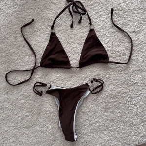 Helt ny & oanvänd Brun bikini - Bikini med trekantsöverdel och knyt i nacken och ryggen. Underdelen har höga skärningar och silverfärgade ringdetaljer på sidorna. Materialet är mjukt och stretchigt. 