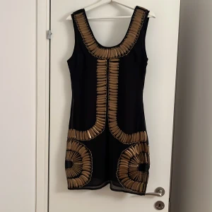 Svart kort klänning med guldmönster - Snygg svart kort klänning med ärmlös design och bred rund halsringning. Klänningen har unika gulddekorationer i geometriska mönster både fram och bak, vilket ger en cool och lyxig vibe. Perfekt för dig som vill sticka ut med stil och detaljer.