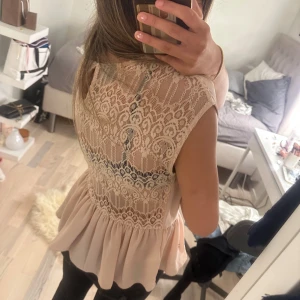 Beige blus med spets och volang - Supersöt beige blus med volang nertill och detaljerad spets på ryggen. Framsidan är enkel och slät medan baksidan har ett vackert spetsmönster som ger en romantisk vibe. Ärmlös modell med rund halsringning och lätt, flowigt tyg.