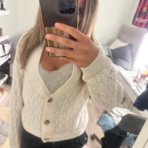 Vit croppad kofta från Mavi - Superfin vit kofta från Mavi med croppad passform och snyggt hålmönster. Tröjan har tre bruna knappar framtill, ribbade muddar och v-ringad hals. Perfekt att slänga över en topp för en chill look. Långärmad och stickad i mjukt material.