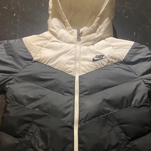 Nike pufferjacka för pojkar för bra pris - (OSÄKER oM STORLEK, DET STÅR PÅ SISTA SLIDEN!) Snygg pufferjacka från Nike i svart och beige med huva och dragkedja framtill. Jackan har ett stilrent blockmönster och klassisk Nike-logga på bröstet. Perfekt för kalla dagar när du vill hålla stilen och värmen.