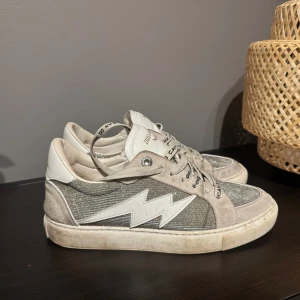 Zadig & Voltaire sneakers med blixtdetalj - Snygga sneakers från Zadig & Voltaire i grått och vitt med glittriga paneler och en vit blixtdetalj på sidan. Skorna har snörning, rund tå och platt sula. Ovandelen är i mocka, skinn och textil, med cool branding på sulan och snörena. Använda men ändå fina tvättas självklart innan köp