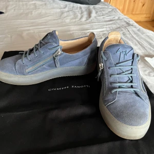 Blå mocka sneakers från Giuseppe Zanotti - Snygga blå sneakers i mocka från Giuseppe Zanotti med coola silverfärgade dragkedjor på sidorna och matchande blå snörning. Skorna har en platt sula och rund tå, samt den ikoniska loggan på plösen. Perfekta för dig som gillar stilrena men unika detaljer. MVH Max🥰