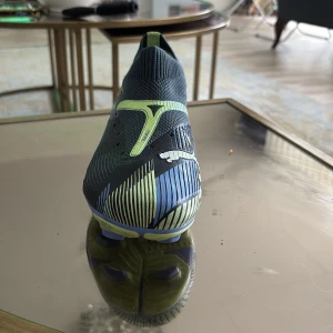 Puma fotbollsskor i blå och neongul - Säljer ett par snygga Puma fotbollsskor i blått med neongula detaljer och vit logga. Skorna har en strumpliknande ovandel och mönstrad sula för bra grepp på planen. Perfekta för dig som vill sticka ut på fotbollsplanen!