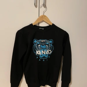 Svart sweatshirt från Kenzo med tigertryck (inte äkta) - Svart sweatshirt från Kenzo med ikoniskt tigertryck i blått och vitt framtill samt texten 'KENZO PARIS'. Tröjan har rund halsringning och långa ärmar. Perfekt för en cool och avslappnad stil. 