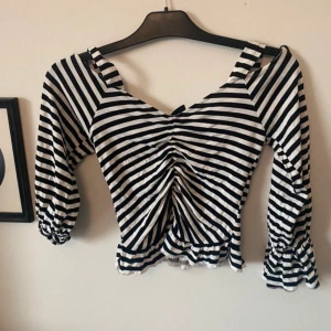 Randig croppad topp från Gina Tricot - Säljer en svartvit randig croppad topp från Gina Tricot med smockade ärmslut och rynkdetalj framtill. Toppen har trekvartsärm och breda axelband. Perfekt för dig som gillar trendiga och unika plagg!Frakt tillkommer